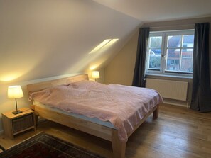 A bedroom with a wooden bed, two bedside lamps, a window with curtains, and a radiator. - Holiday Apartment With 2 Bedrooms (Barkelsby)
