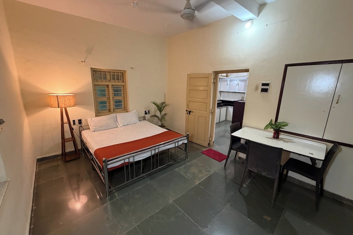 A bedroom with a bed, bedside table, lamp, and a view into the kitchen.