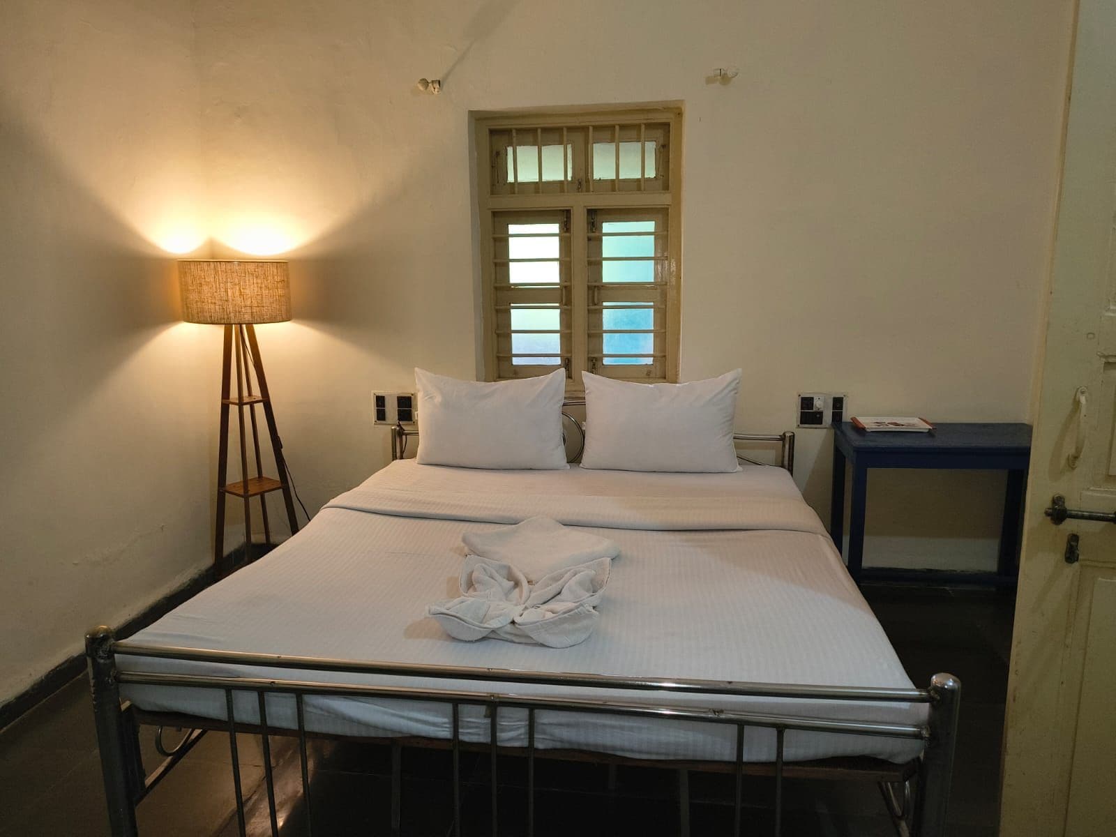 Un lit simple avec un cadre en métal, une literie blanche et une lampe de chevet. Il y a une fenêtre avec des volets en bois et une petite table avec une chaise sur la droite.
