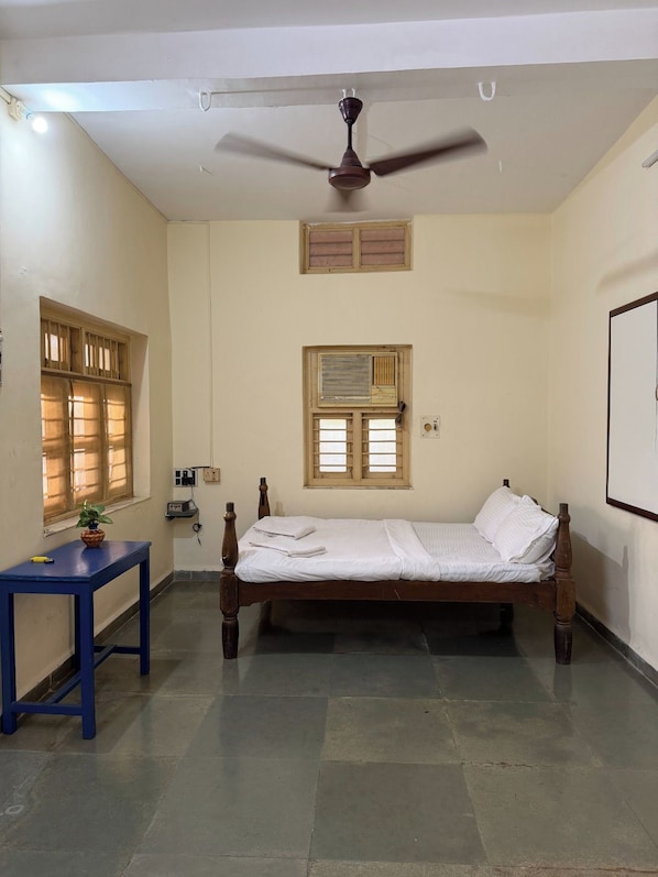 A room with a bed, a table, a ceiling fan, and two windows.