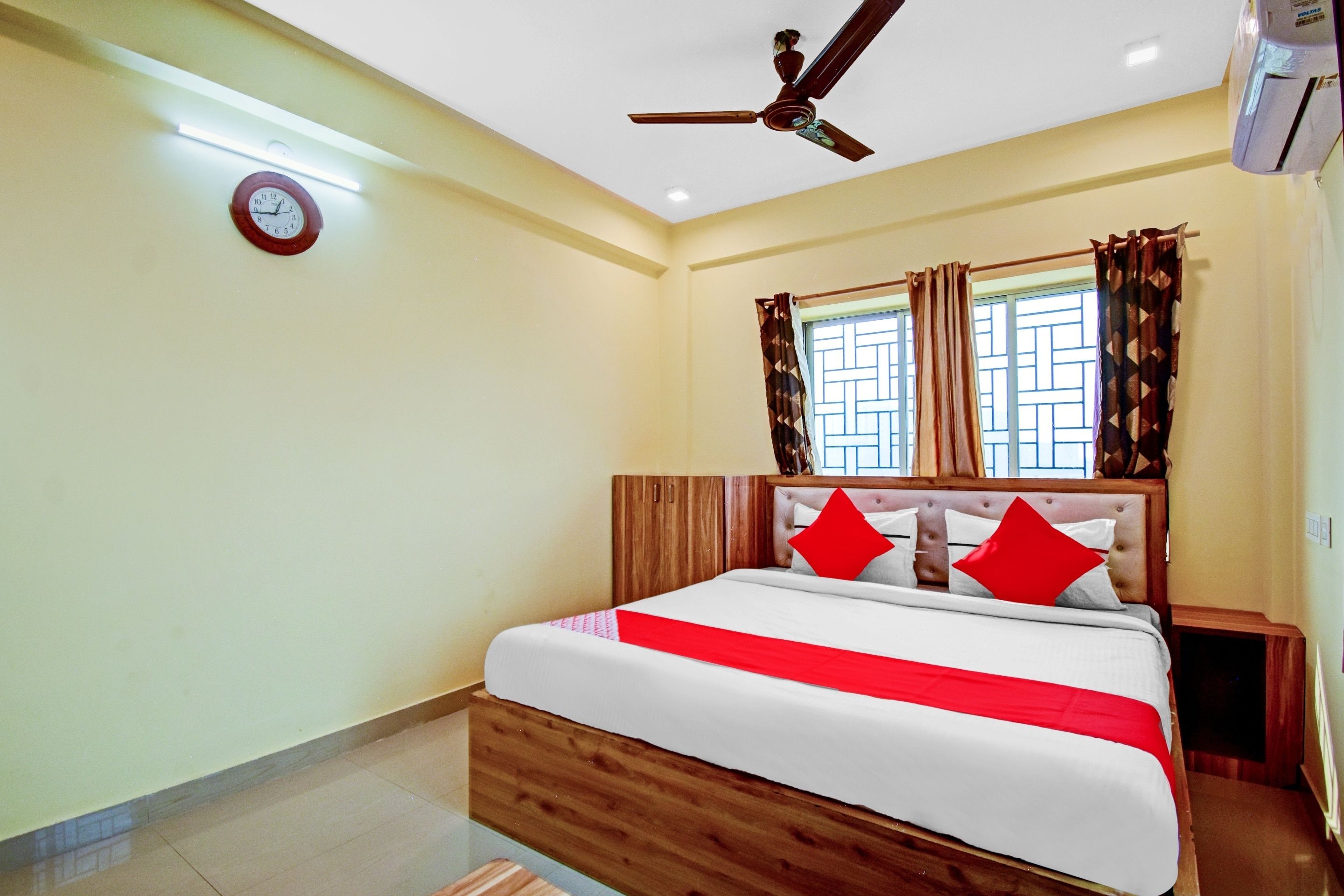 A hotel room with a bed, a ceiling fan, a clock, and a window with curtains.