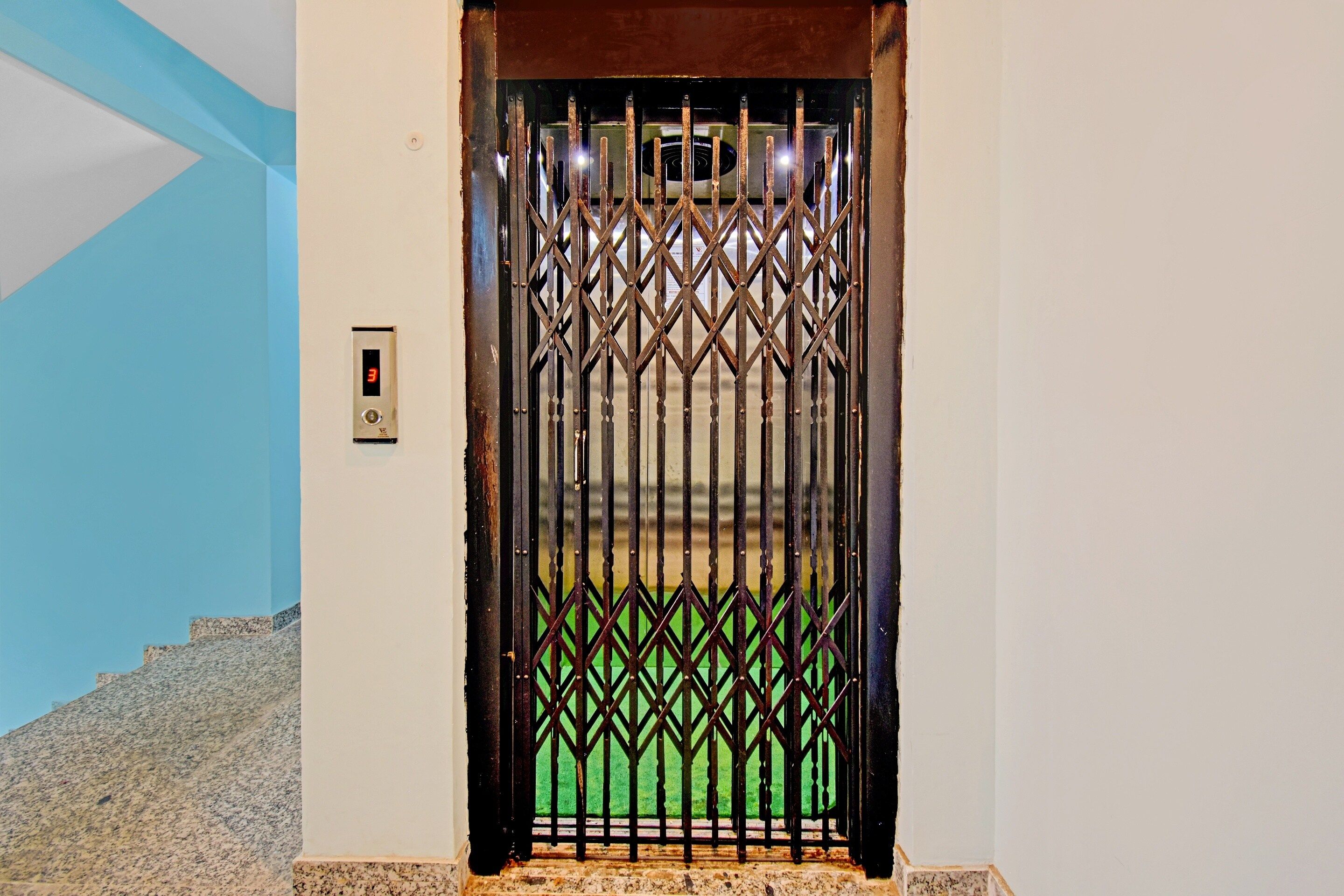Un ascensor con una puerta metálica, un panel digital y un suelo verde visible a través de la puerta.