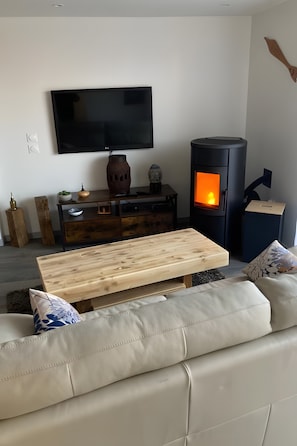 Een woonkamer met een bank, een houten salontafel, een aan de muur bevestigde televisie en een houtkachel.