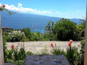 A table with two wine glasses, surrounded by greenery and flowers, overlooking a mountainous landscape. - Casa Orio Apt. 2 (Tignale)