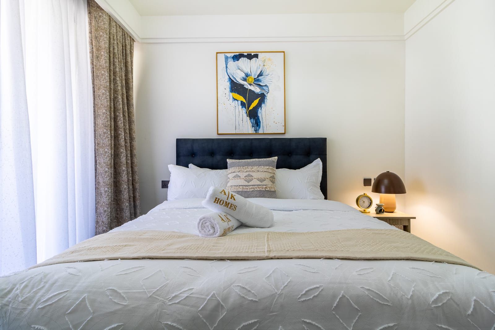 Una habitación de hotel con cama, un cuadro sobre la cama, una mesita de noche con lámpara y una ventana con cortinas.