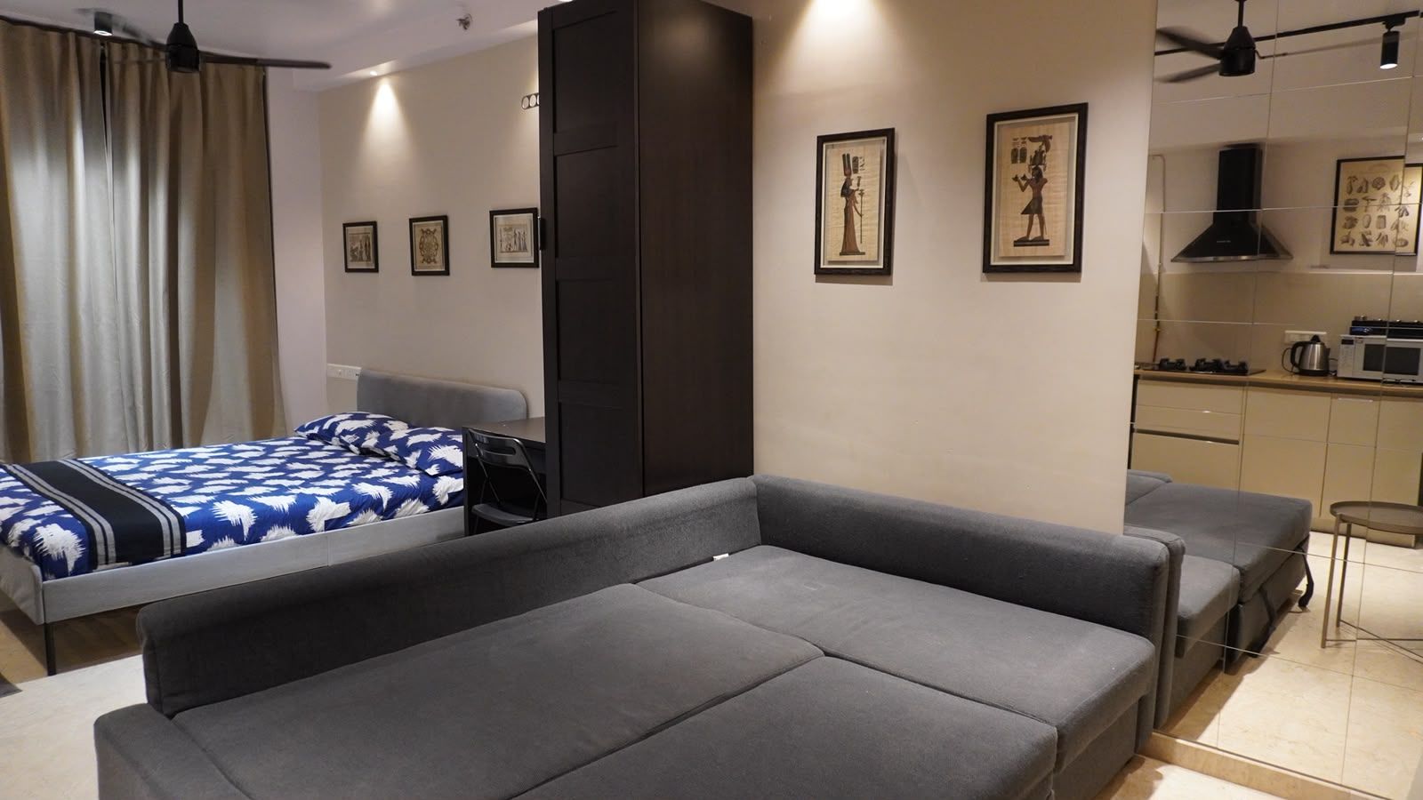 A modern hotel room with a sofa, a bed with a patterned bedspread, a desk, a chair, and a kitchenette area.