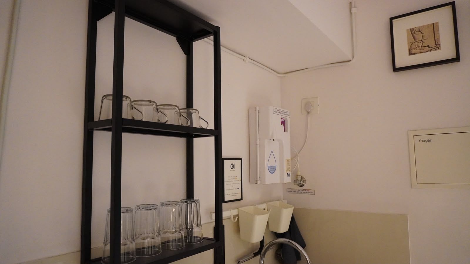 A room with a black metal shelf holding clear glasses, a white water dispenser, and a framed picture on the wall.