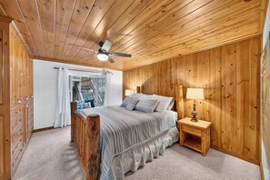 A bedroom with a wooden ceiling, a bed with a striped bedspread, a nightstand with a lamp, and a view of a deck through a window. - Lakefront Retreat • Boat Dock + Hot Tub | Near Beach | Meeks Bay Chalet (Tahoma)