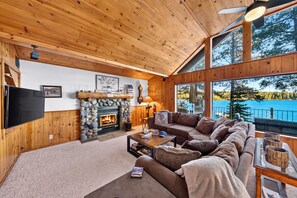 A cozy living room with a stone fireplace, wooden walls, a large window overlooking a lake, and a flat-screen TV mounted on the wall. - Lakefront Retreat • Boat Dock + Hot Tub | Near Beach | Meeks Bay Chalet (Tahoma)