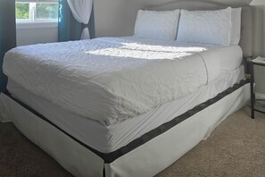 Una cama bien tendida con cubrecama y almohadas blancas, una ventana con cortinas y una mesita de noche pequeña.