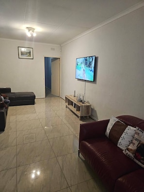 Ein Wohnzimmer mit an der Wand montiertem Fernseher, einem Sofa, einem Couchtisch und einer Tür zu einem anderen Raum.