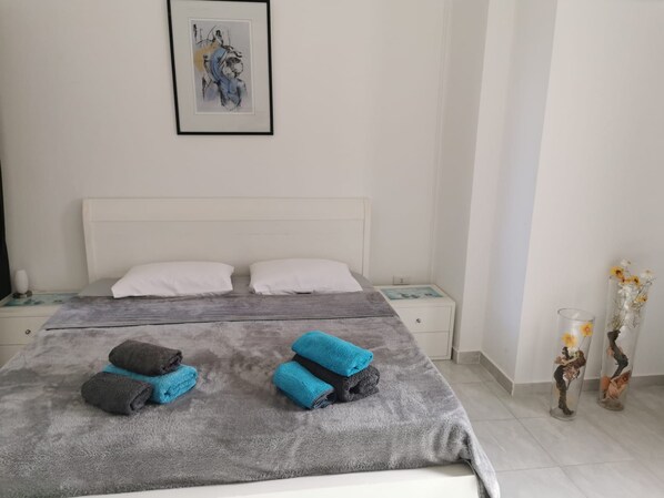 Een opgemaakt bed met een grijze deken en twee kussens, een nachtkastje met een lamp, en een lijst met een kunstwerk aan de muur.