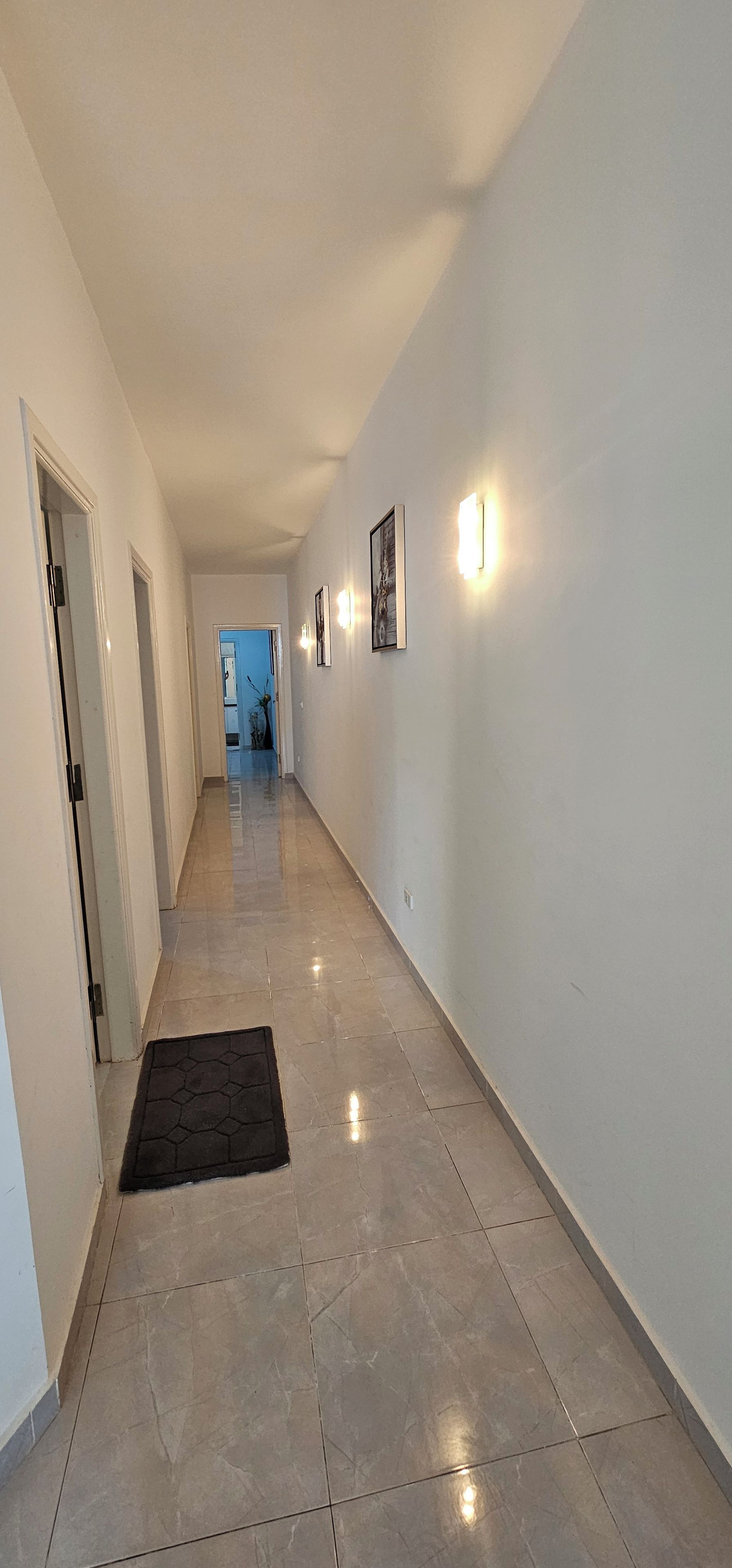A hallway with a tiled floor, a black doormat, and framed pictures on the wall.