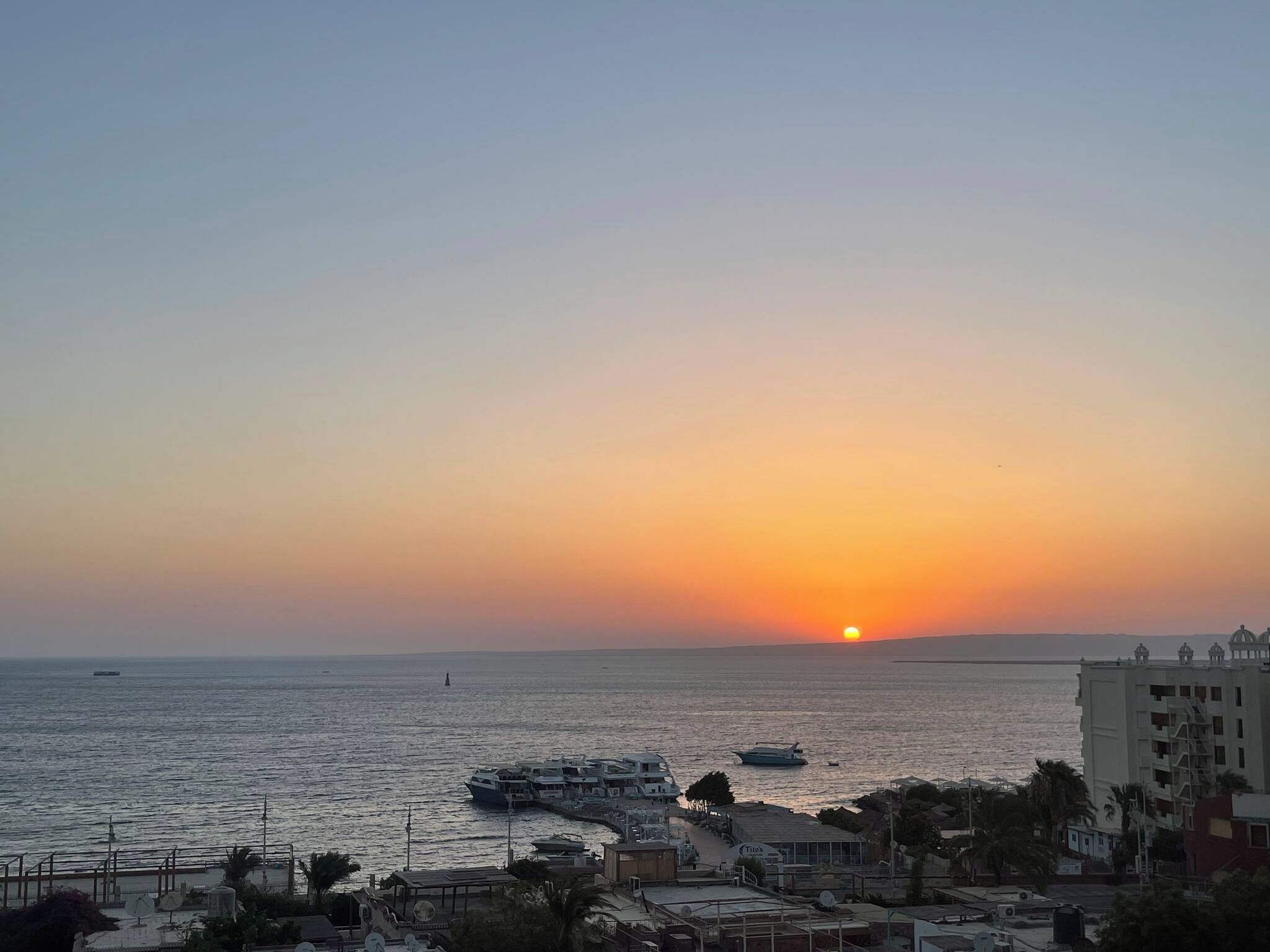 A coastal sunset view with boats, buildings, and a clear sky.