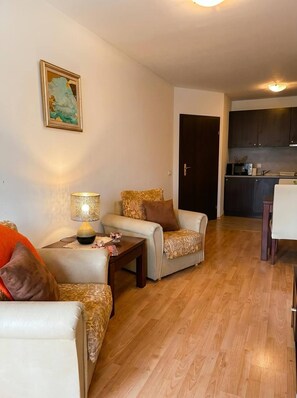 A living room with a sofa, armchair, and coffee table, a dining table with chairs, a kitchen area, and a door. - South Facing Balcony 1 BR (Bansko)