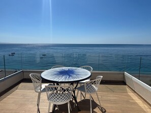 Une table ronde ornée d’un motif bleu, entourée de chaises blanches, donnant sur une mer calme où voguent des bateaux.