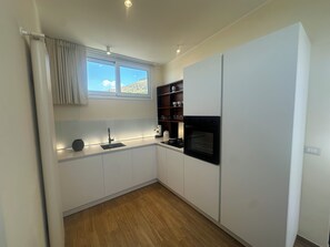A compact kitchen with white cabinets, a black oven, and a window with curtains. - Blue Wave Apartment: Relax by the Sea, Panoramic Terrace, and Modern Design (Letojanni)