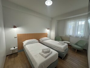 A hotel room with two beds, a small table, a chair, and a window with curtains. - Blue Wave Apartment: Relax by the Sea, Panoramic Terrace, and Modern Design (Letojanni)