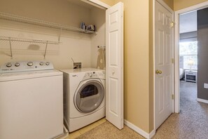 A laundry room with a front-loading washer and dryer, a shelf above the washer, and a door leading to another room. - Family Townhome Near Disney | Pool | Gated (Kissimmee)