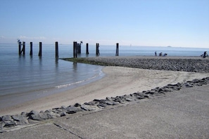 Ein Strand mit einer Betonpromenade, Kieselsteinen und Holzpfählen im Wasser.