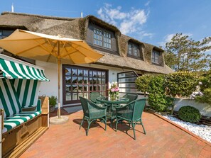 A thatched-roof house with a patio area featuring a table and chairs, a striped awning, and a window with a view. - Holiday Apartment Oystercatcher (Kampen)