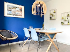 A dining area with a round table, chairs, a wicker chair, a wall-mounted light fixture, and a framed collage. - We05 Holiday Home With Outdoor Pool (Westkapelle)