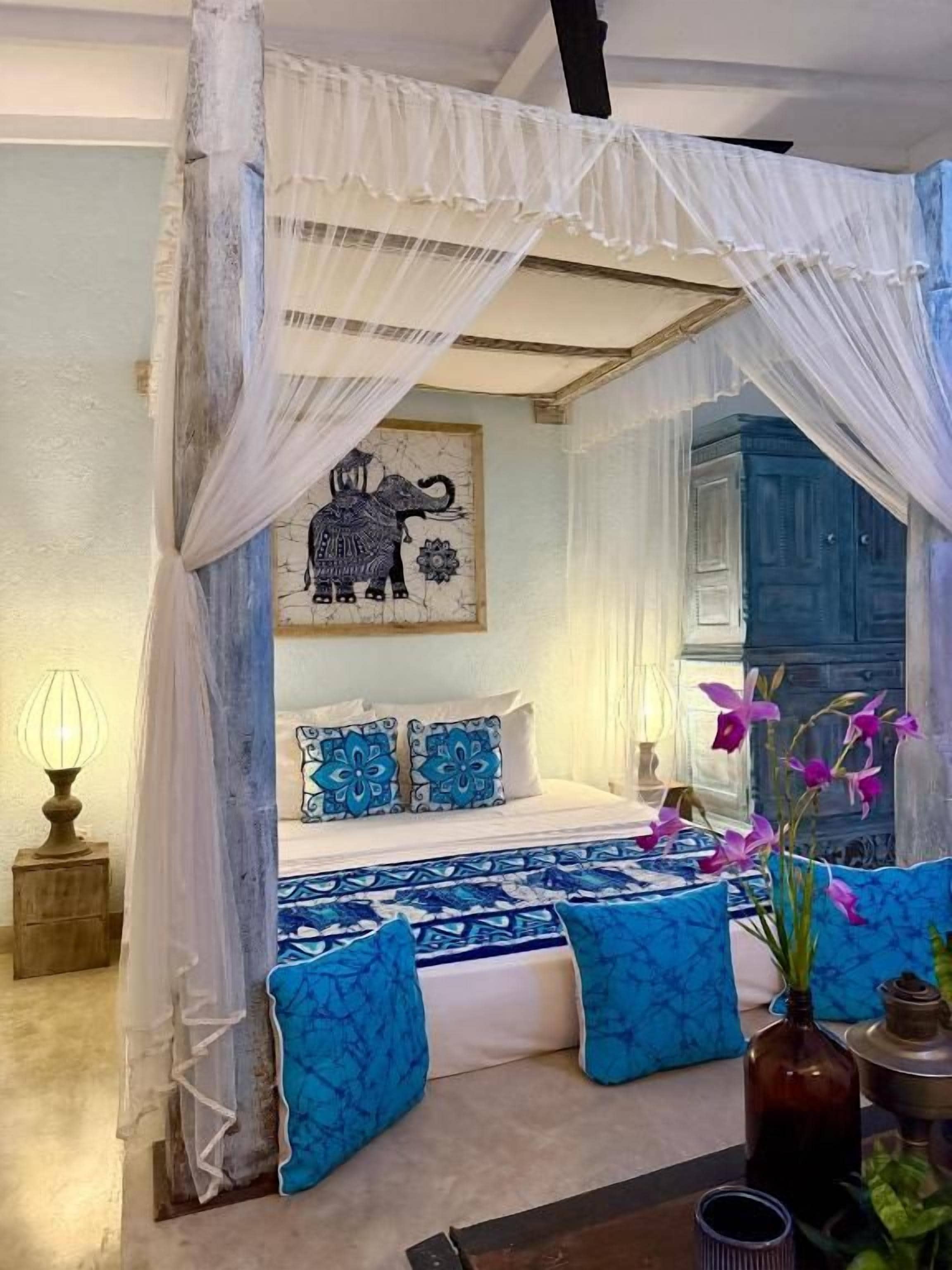 Ein Schlafzimmer mit Himmelbett, blau gemusterten Textilien, einer Blumenvase und einem Wandbild.