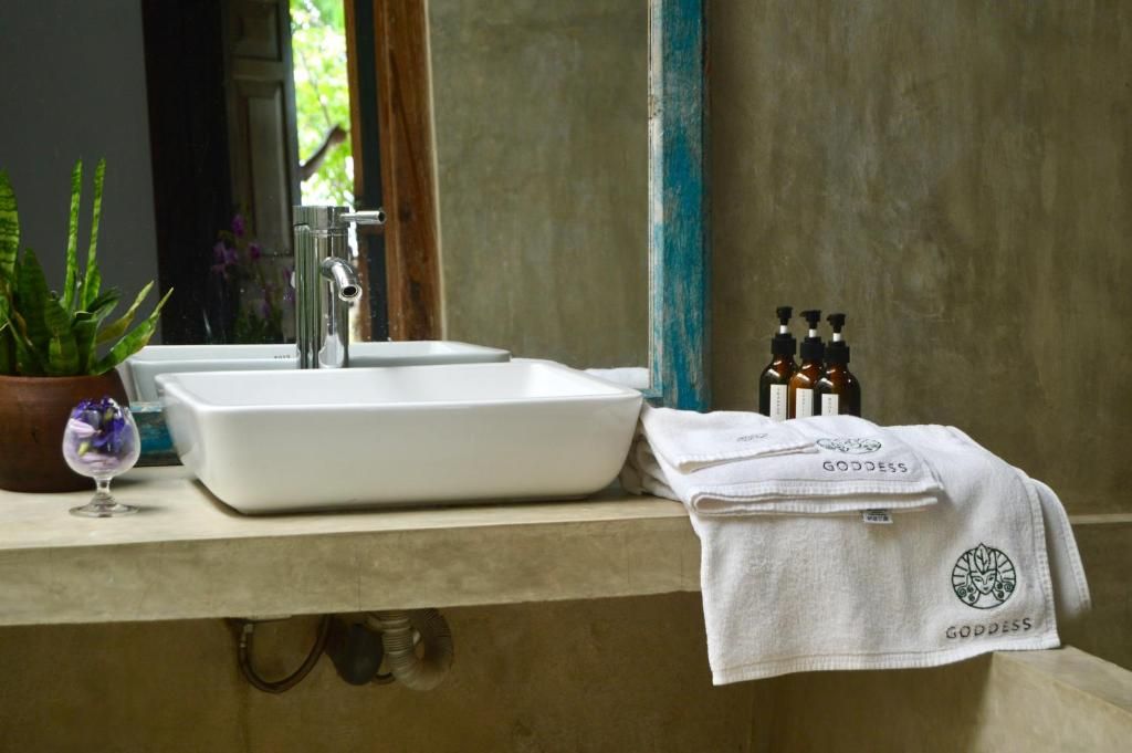 Une salle de bain avec un lavabo blanc, un miroir et une serviette ornée d’une broderie représentant le mot « DÉESSE ».