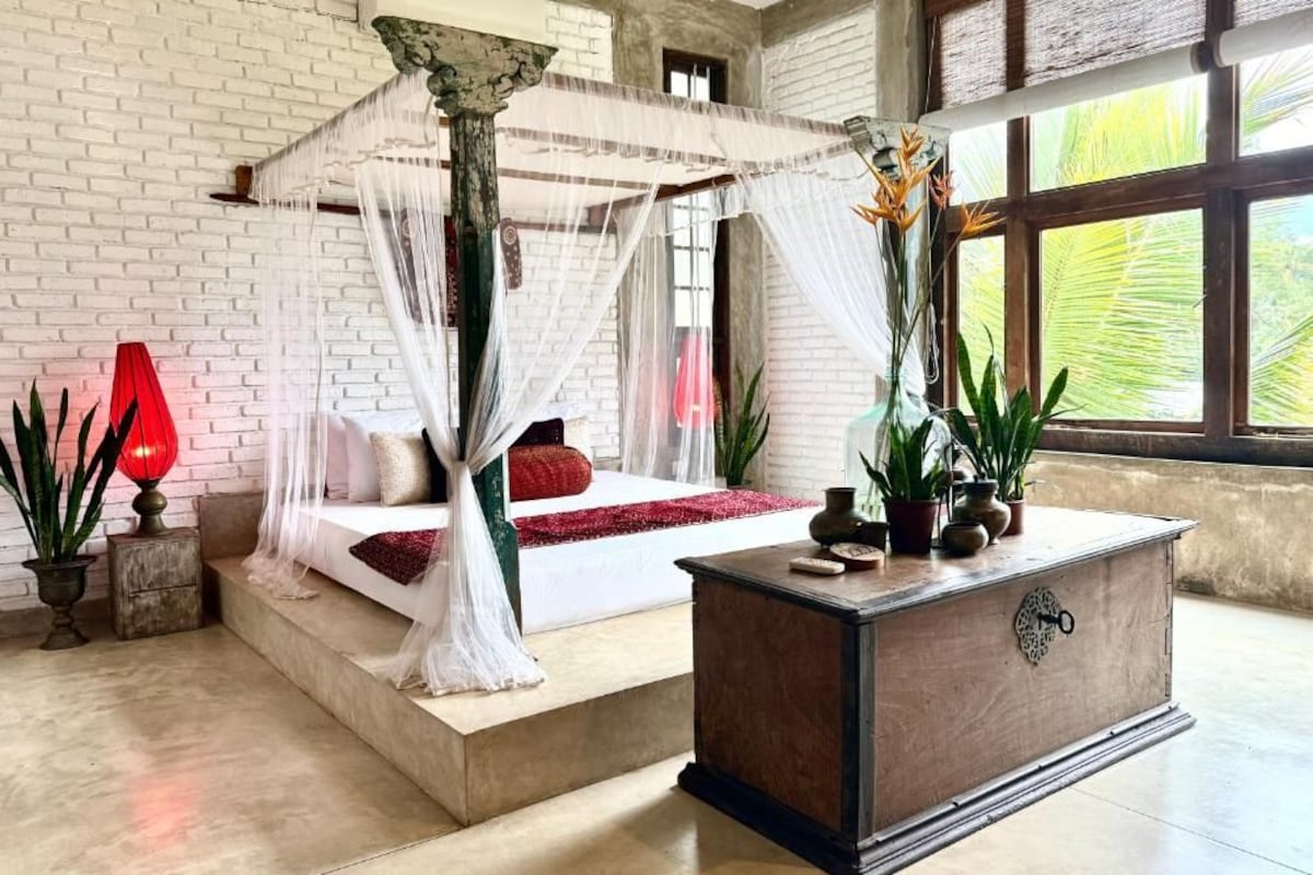 Un dormitorio con una cama con dosel, una mesita de noche de madera y un ventanal con vistas a la vegetación.