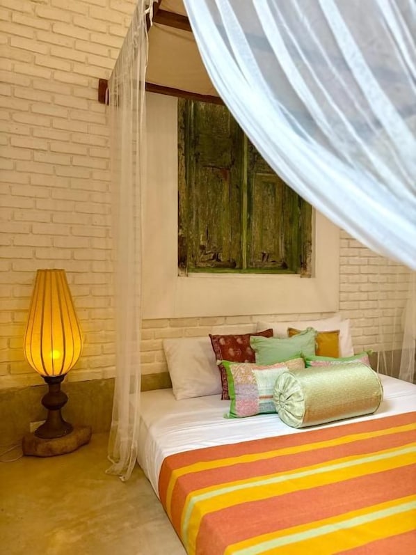 A bedroom with a bed, a lamp, a window with curtains, and a brick wall.