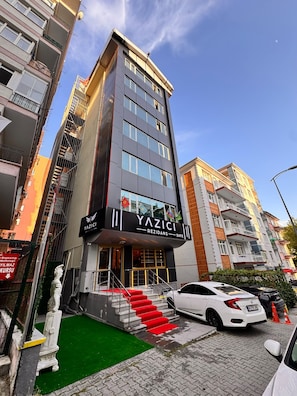 A multi-story building with a prominent entrance, a staircase with red carpet, and a white car parked in front. - YAZICI APART REZİDANS YENİMAHALLE (Ankara)