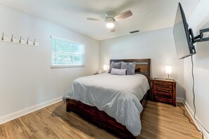 Ein Schlafzimmer mit einem Bett, einem Nachttisch, einem Deckenventilator, einem Fenster mit Jalousien und einem an der Wand montierten Fernseher.