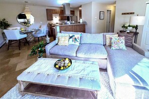 A living room with a sofa, a coffee table with a bowl of fruit, a dining area with a table and chairs, and a kitchen in the background. - Flatiron Views, Pool Access! Boulder Extended Stay (Boulder)