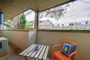 A covered outdoor seating area with a table and a chair, a view of a residential building, and a metal fence with signs. - Flatiron Views, Pool Access! Boulder Extended Stay (Boulder)