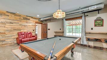 Une table de billard dans une pièce dont un mur est en bois, avec un canapé en cuir rouge et un lustre.