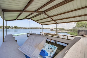 A covered dock area with a boat, a table, and a view of the water and distant buildings. - San Angelo Lake Escape: Dock & Fire Pit (San Angelo)