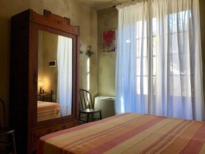 Ein Schlafzimmer mit einem großen Spiegel, einem Bett mit gestreifter Bettwäsche, einem Sessel und einem Fenster mit transparenten Vorhängen.