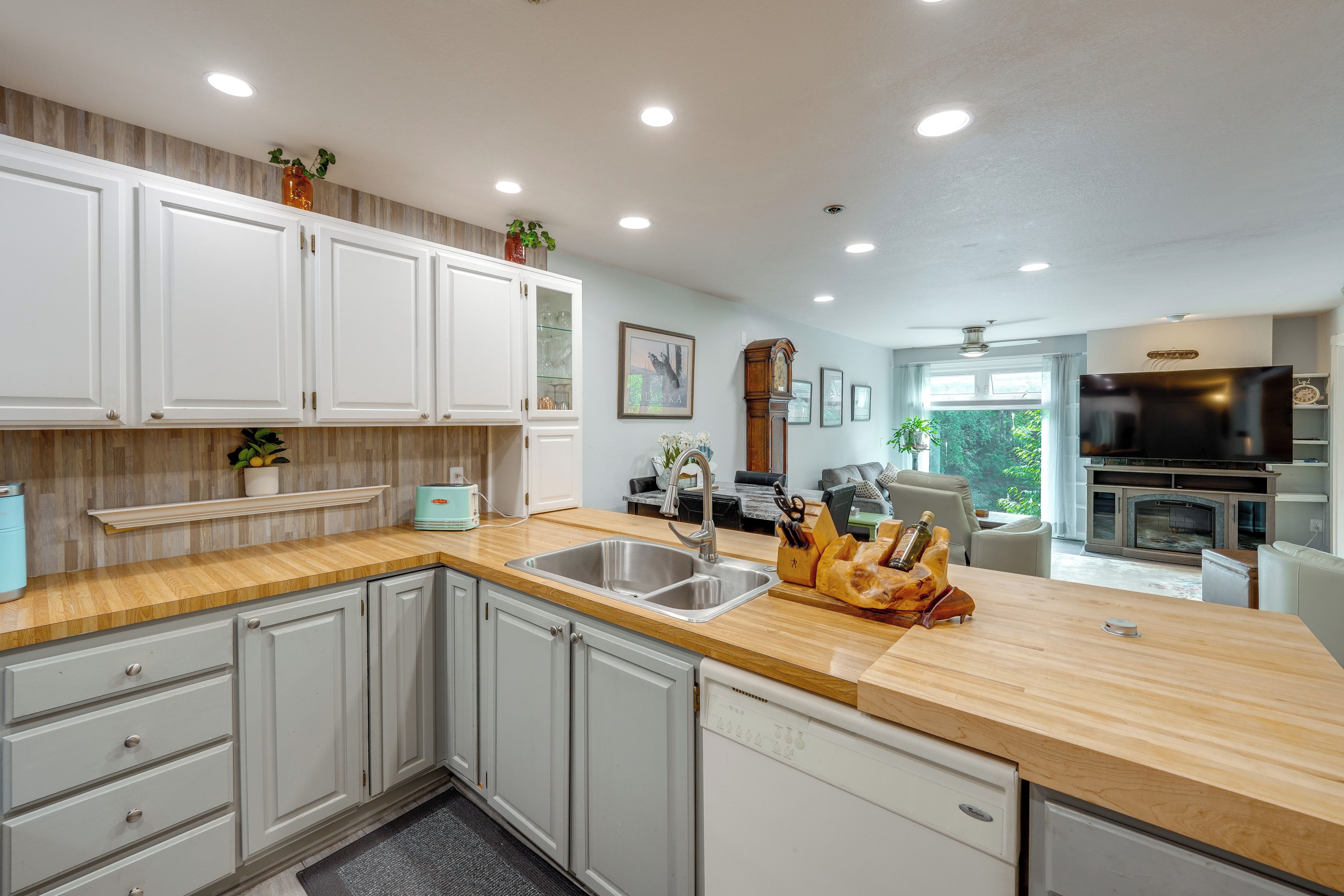 A modern kitchen with white cabinets, a wooden countertop, a built-in dishwasher, and a large window providing natural light.