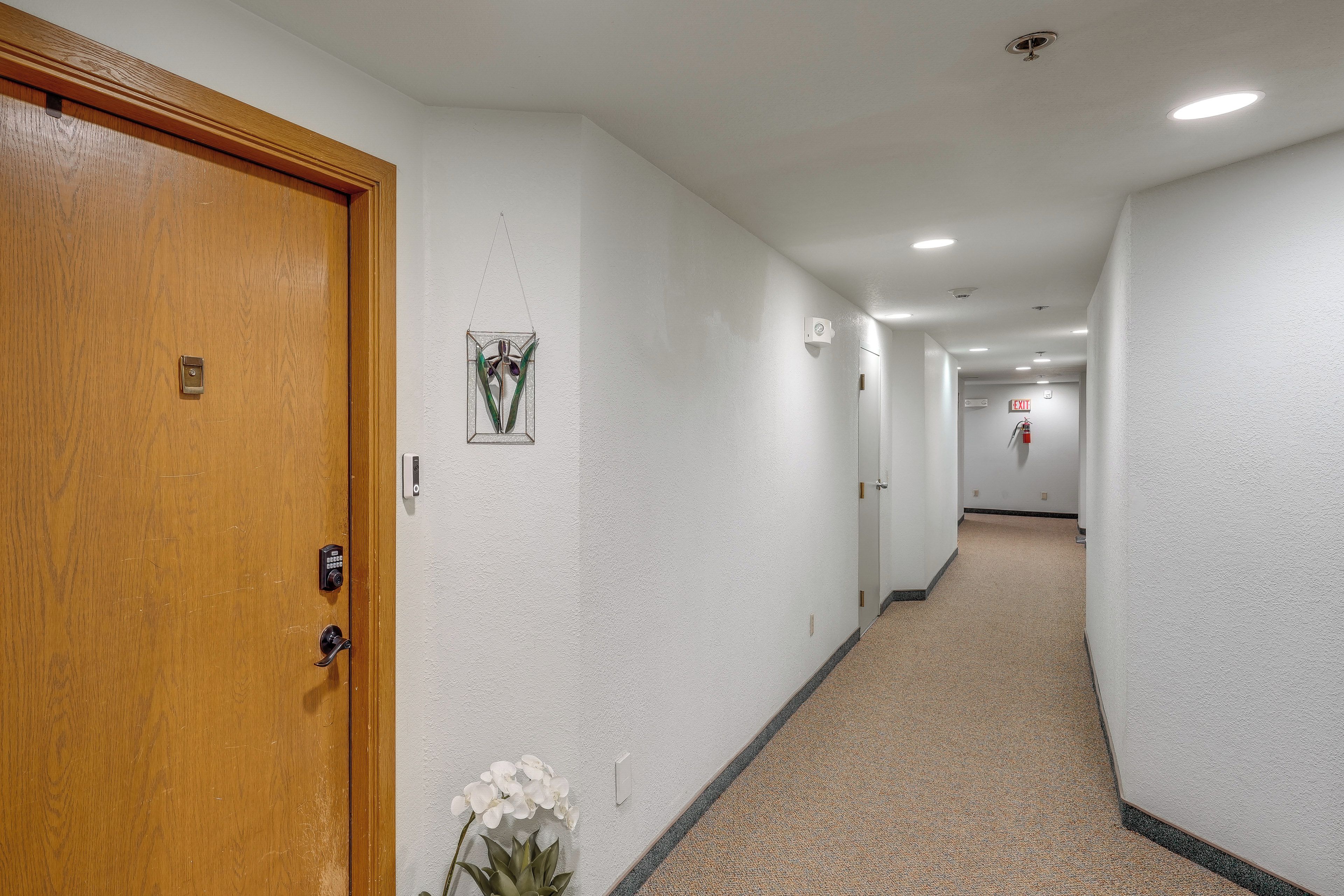 A hotel hallway with a wooden door, a security camera, a fire alarm, and a light switch on the wall.