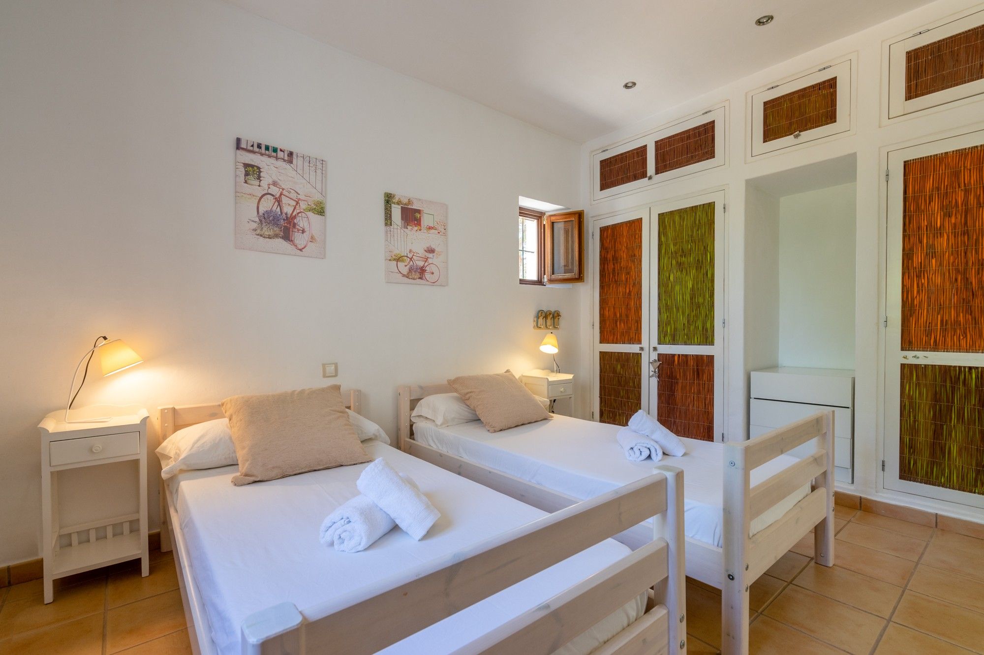 Habitación de hotel con dos camas, una mesita de noche, una lámpara y una ventana con persianas de madera.