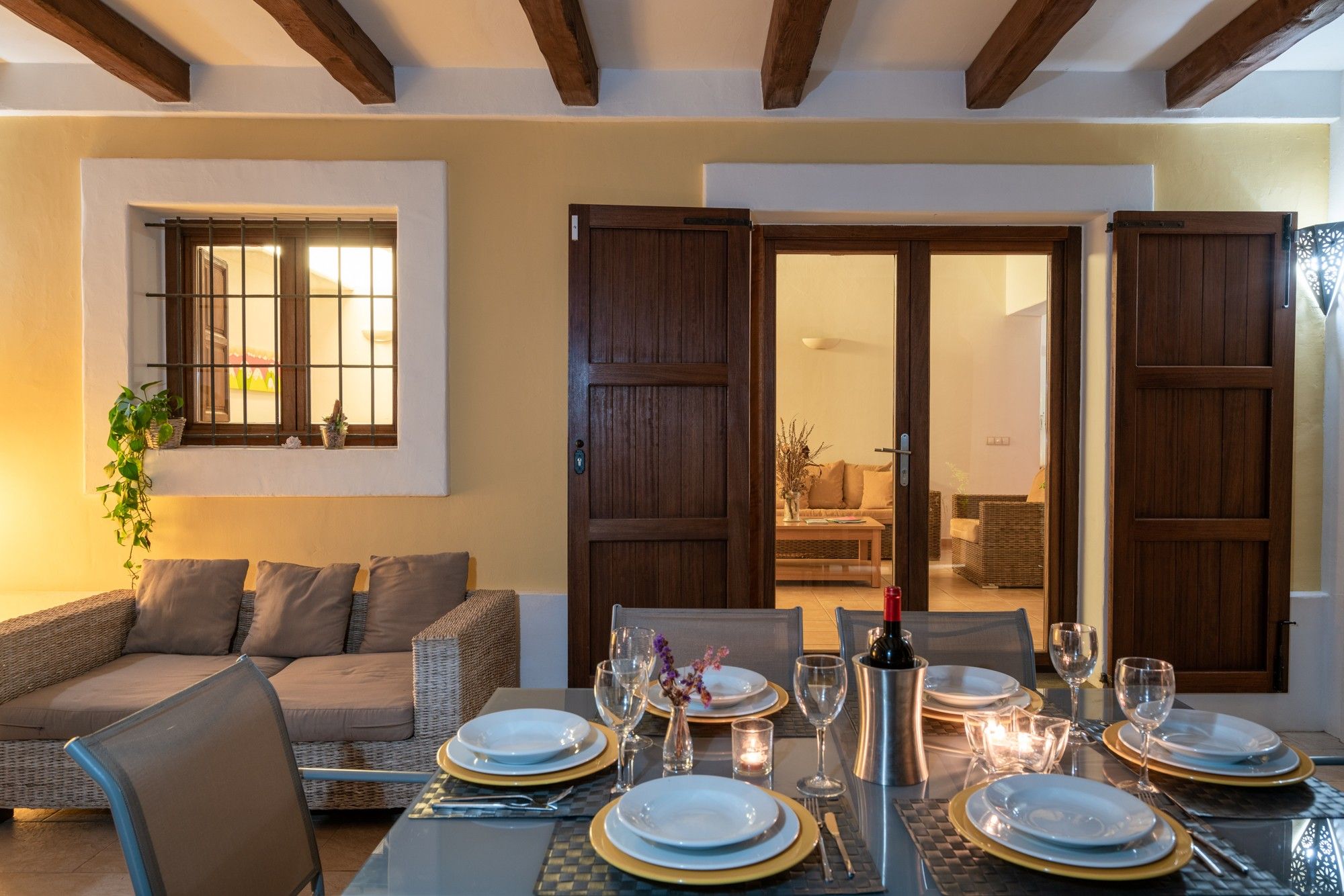 Un comedor con una mesa preparada para cuatro personas, un sofá y una ventana con plantas.