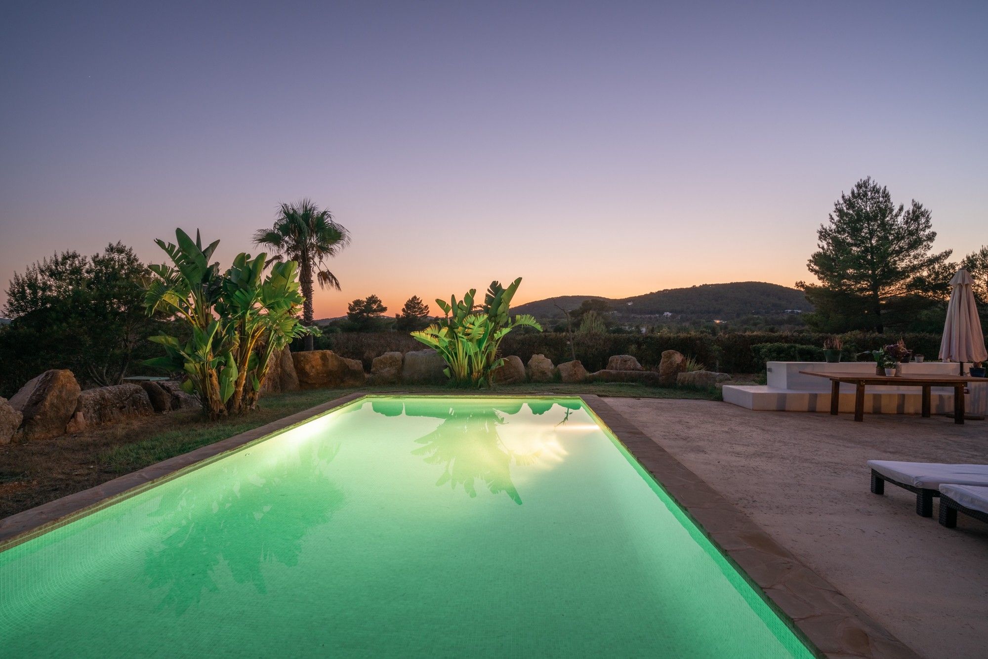 Una piscina rectangular con borde transparente, rodeada de rocas y plantas, con vistas a un paisaje montañoso al fondo.