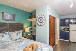 Ein Schlafzimmer mit einem Bett, einem Nachttisch, einer Wanduhr und Blick in eine Küche.