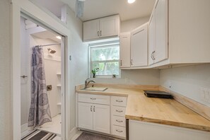 Cozinha compacta com armários brancos, bancada de madeira, janela com vista para o verde e área de chuveiro com cortinas estampadas.
