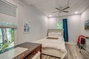 Quarto com cama, escrivaninha, ventoinha no teto, janela com persianas e um quadro na parede.