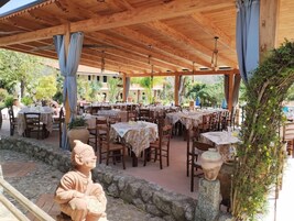 Un espacio para comer al aire libre con varias mesas y sillas, cubierto por una pérgola de madera y rodeado de muros de piedra y follaje.