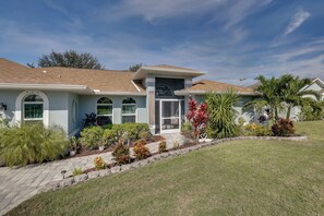 A two-story house with a covered front porch, a garage, and a landscaped front yard with various plants and a lawn. - Saltwater Pool & Firepits: Cape Coral Home (Cape Coral)