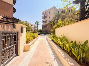 A pathway flanked by a gated entrance and a wall with greenery, leading to a building with multiple windows and balconies.