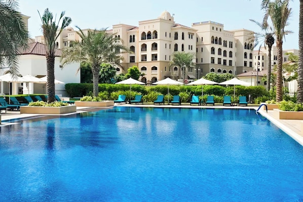A swimming pool with lounge chairs and umbrellas, surrounded by palm trees and a large building with arches.