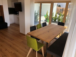 Ein Essbereich mit Holztisch und Stühlen, einem Fernseher an der Wand und Blick ins Freie durch große Fenster.